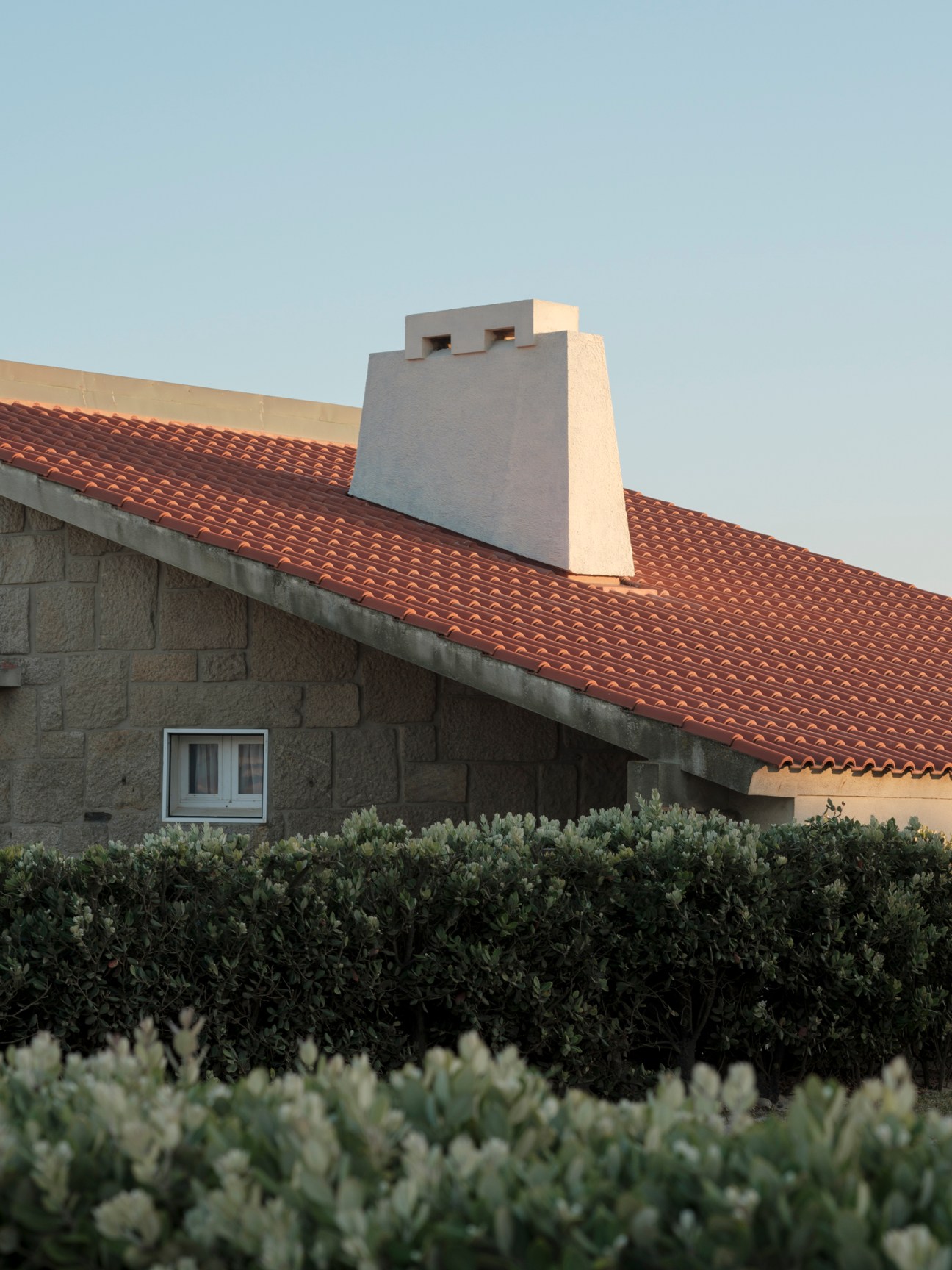 Modern house with red clay tile roof, white chimney, stone walls, and landscaped hedges