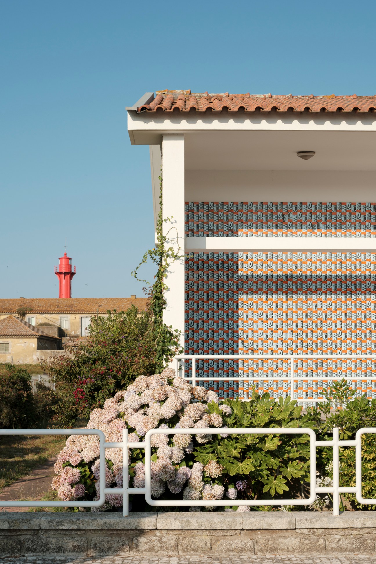 Modern house with patterned tile facade, terra cotta roof, white railings and flowering plants in foreground