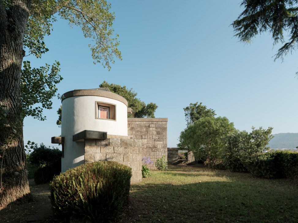 White cylindrical windmill structure with small window attached to stone wall surrounded by trees and grass