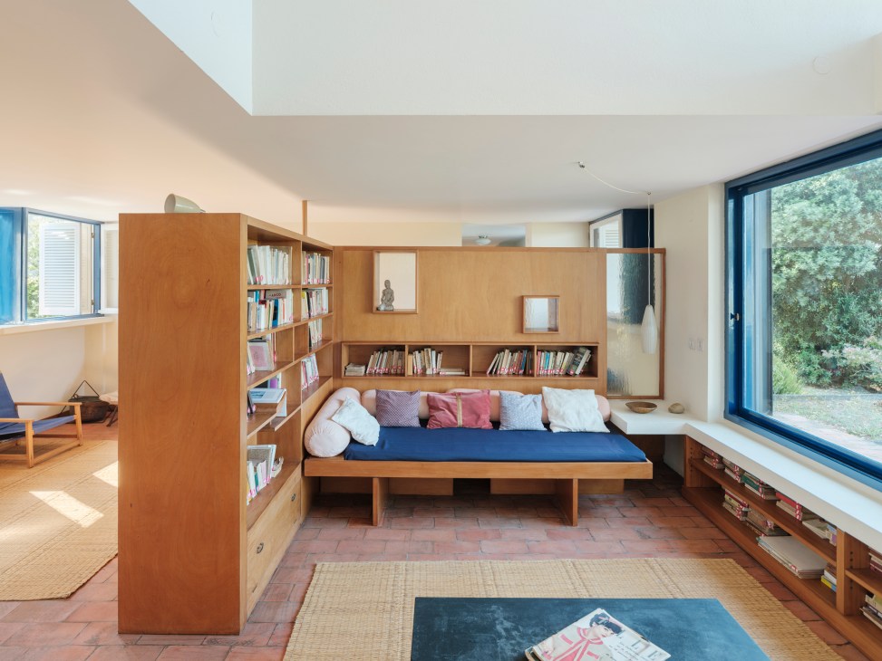 Open-plan living room with built-in wooden bookshelves, daybed with pillows, and large windows overlooking greenery