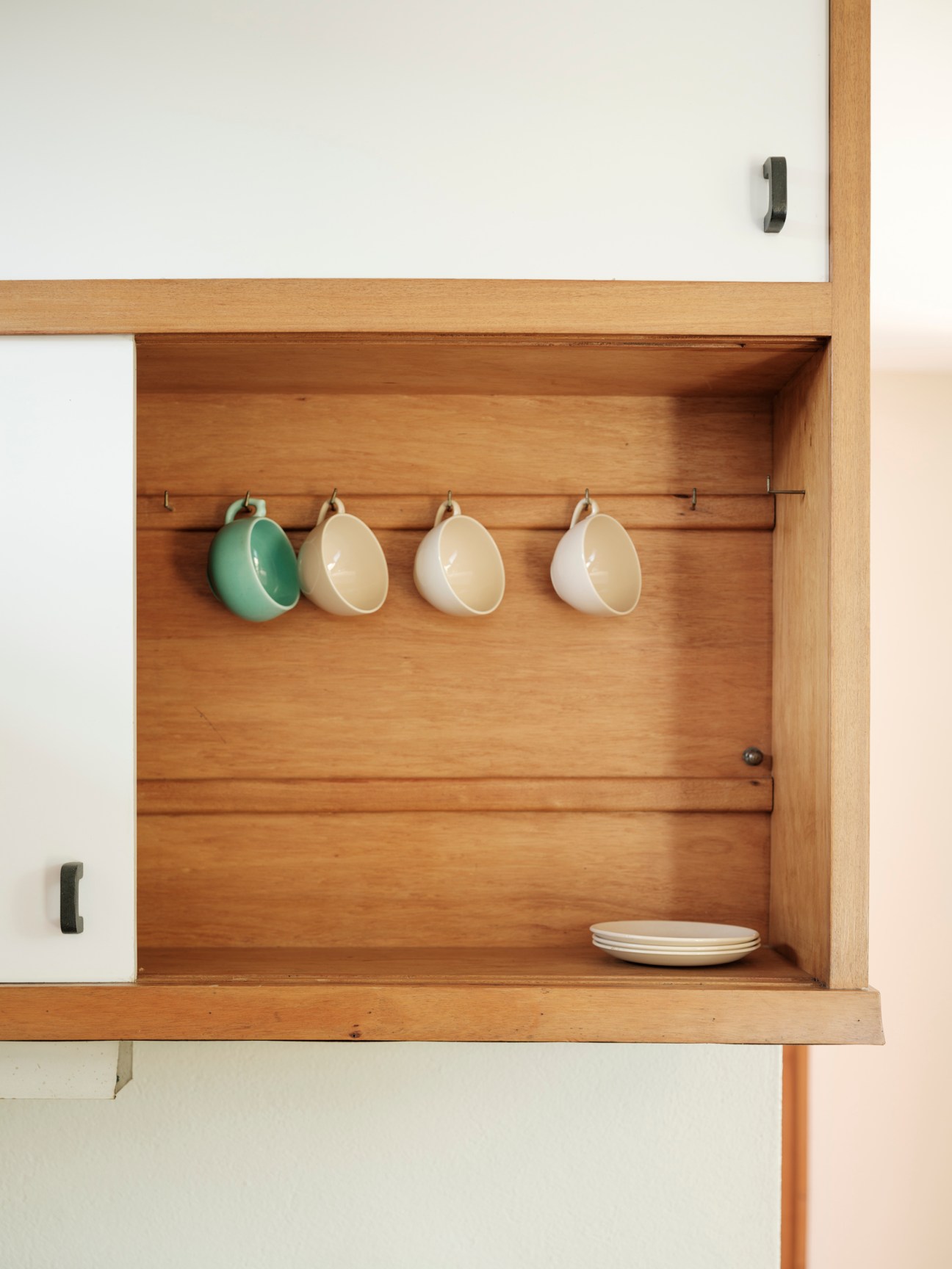 Four ceramic mugs hanging from hooks on wooden rod inside open cabinet with white plates on shelf below