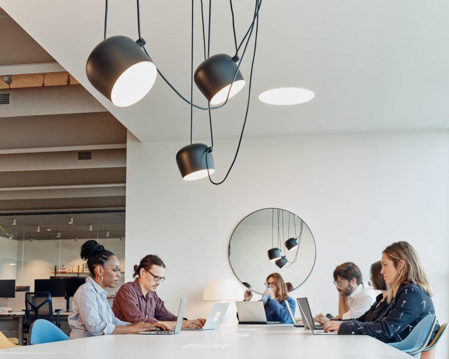 Group of people working on laptops around white table in modern office with pendant lighting and circular mirror