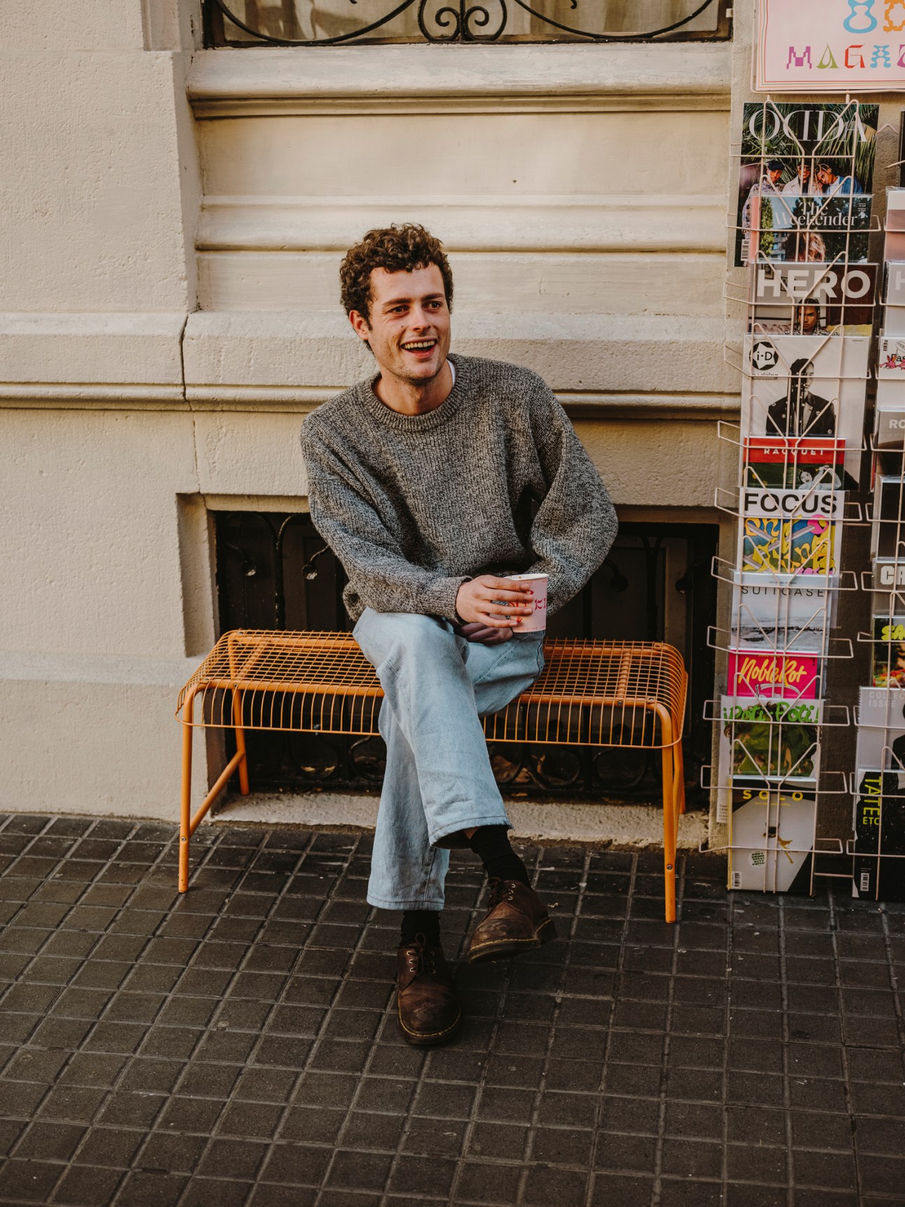 Young man with curly hair sitting on orange bench outdoors next to magazine rack, smiling