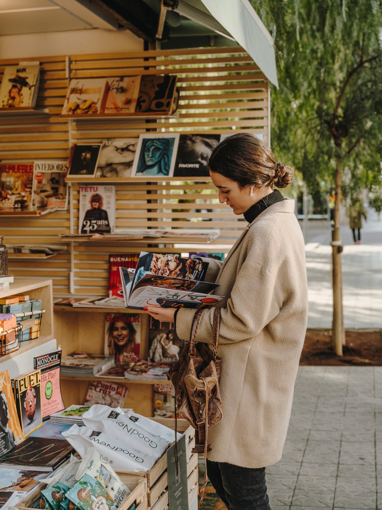 Woman in beige coat reading magazine at outdoor newsstand with books and publications displayed