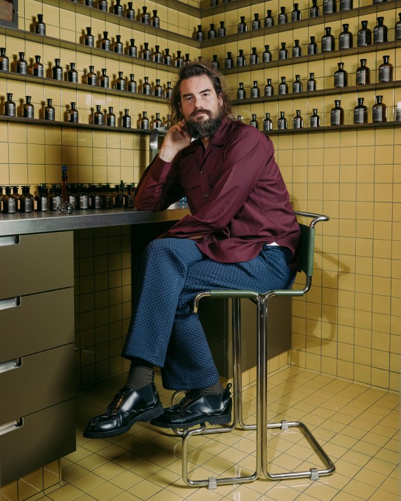 Barnabé Fillion, founder of Arpa fragrance brand, sits in front of shelves of dark glass bottles in a yellow-tiled room.