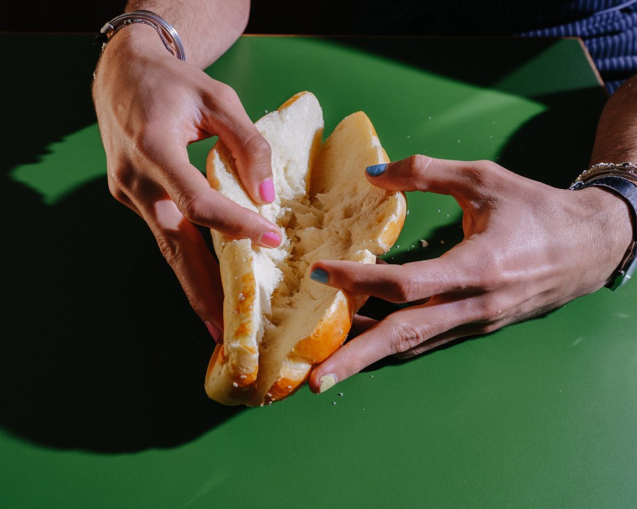 Hands tearing apart fluffy challah bread filled with Mediterranean ingredients on a green surface