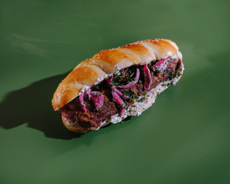 A sandwich with meat, pickled onions and herbs on a challah bun against a green background