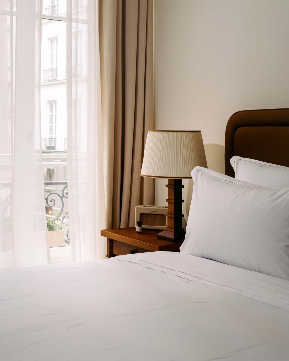 A bright hotel guest room with white bedding, dark brown headboard, wooden nightstand with table lamp, and floor-to-ceiling window overlooking a Parisian street.