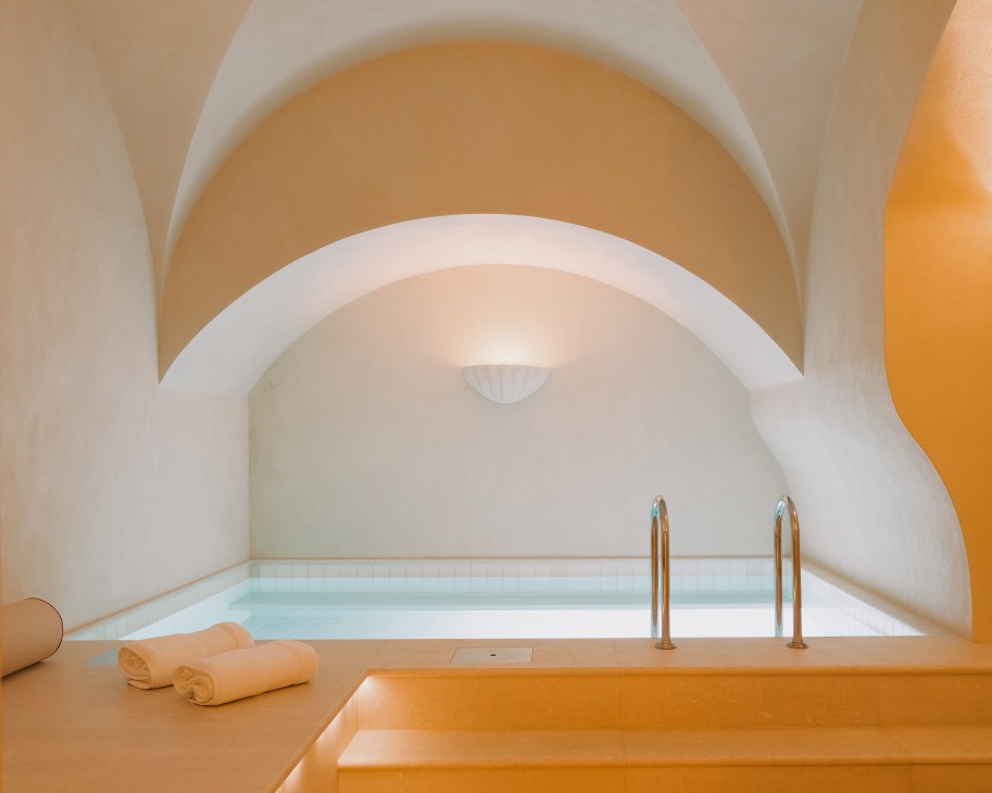 A minimalist spa pool at Château Voltaire with arched white ceiling, golden walls, and rolled towels.