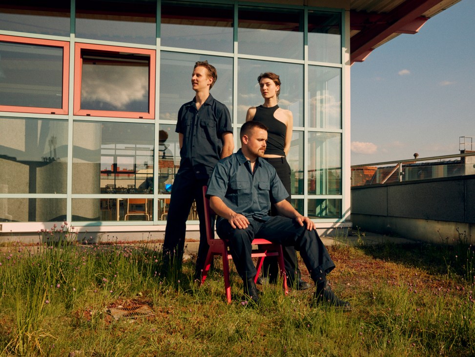 Three people pose on a rooftop terrace with glass windows and urban buildings in the background during golden hour