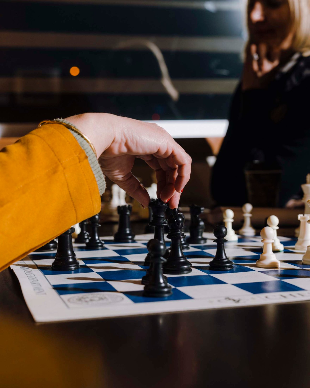 Person in yellow jacket makes a chess move on a blue and white chessboard.
