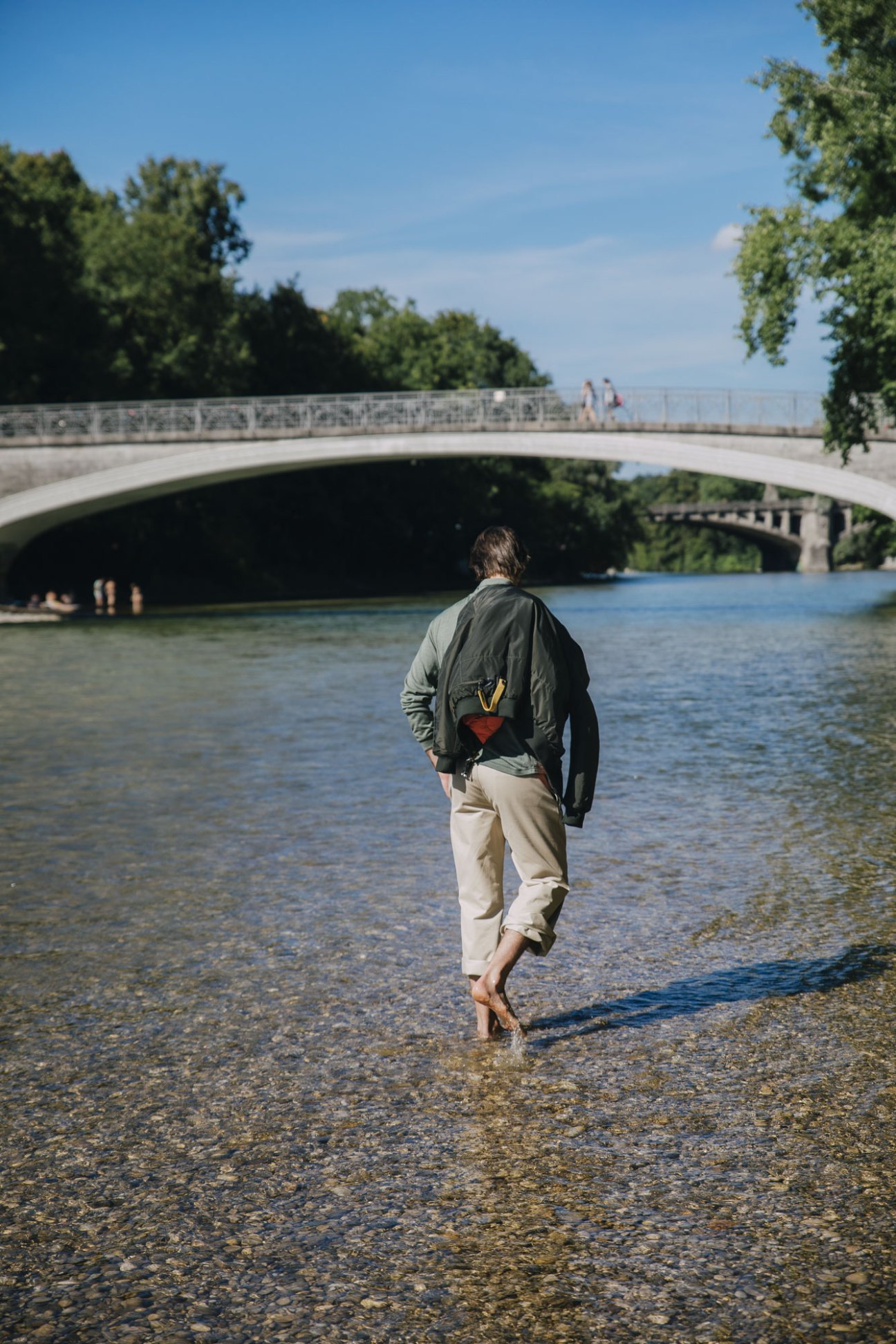 Person wading barefoot through shallow river water beneath an arched stone bridge on a sunny day