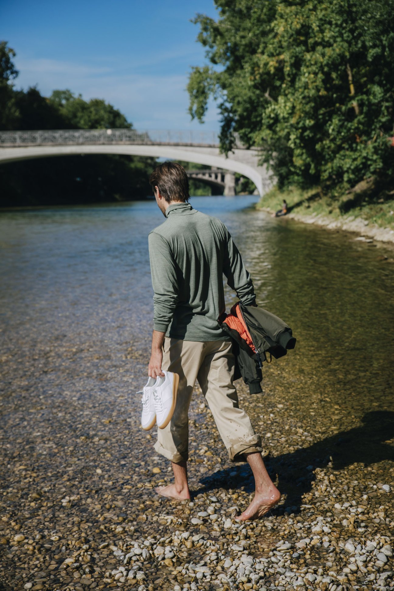 Man walking barefoot on riverbank carrying white sneakers and jacket with stone bridge in background