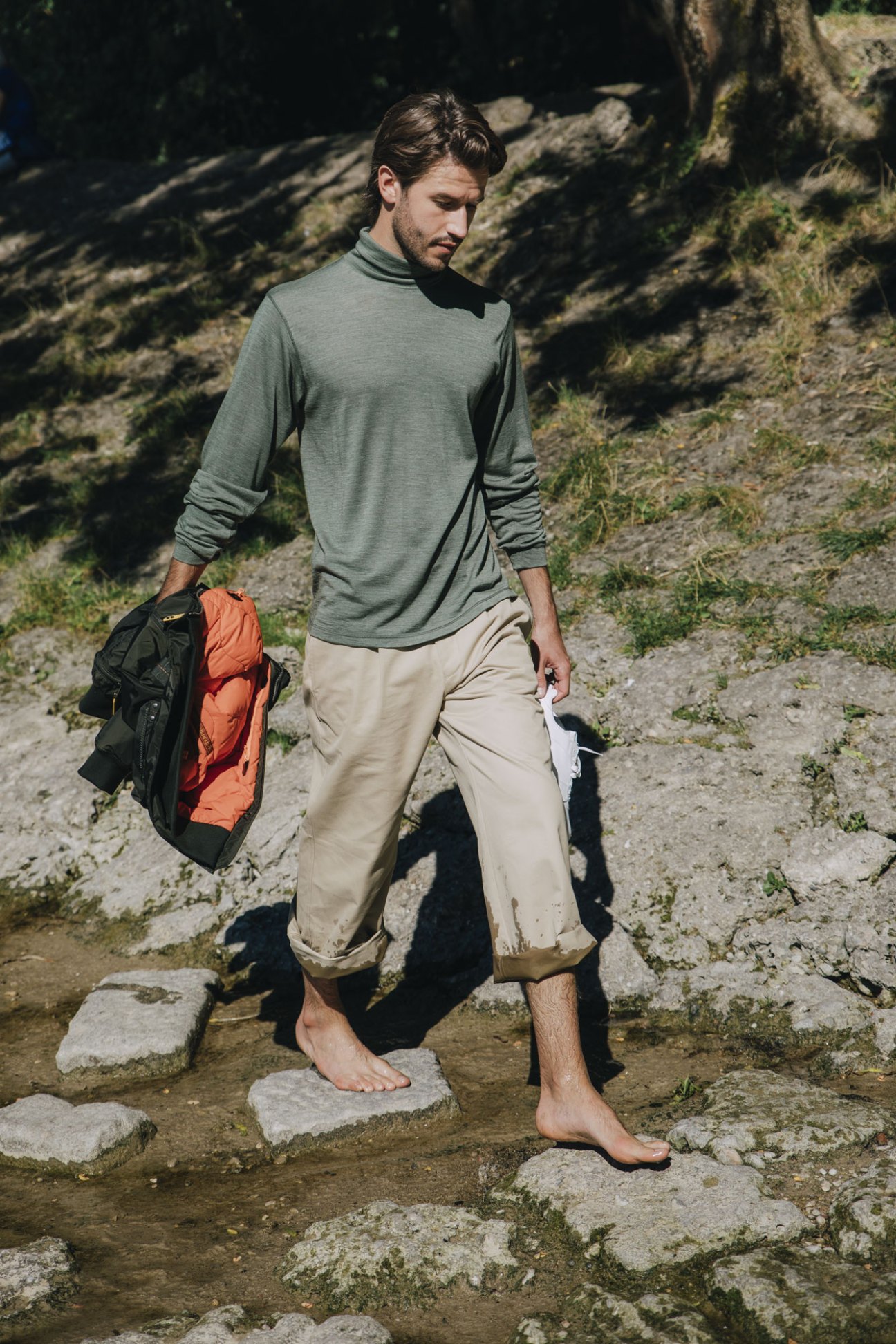 Man in sage green shirt and beige pants walking barefoot on stone path, carrying dark jacket with orange lining