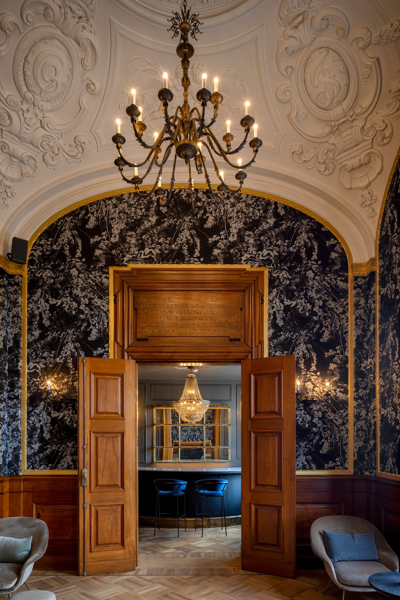 Ornate meeting room with elaborate ceiling moldings, chandelier, dark patterned wallpaper and wooden doors