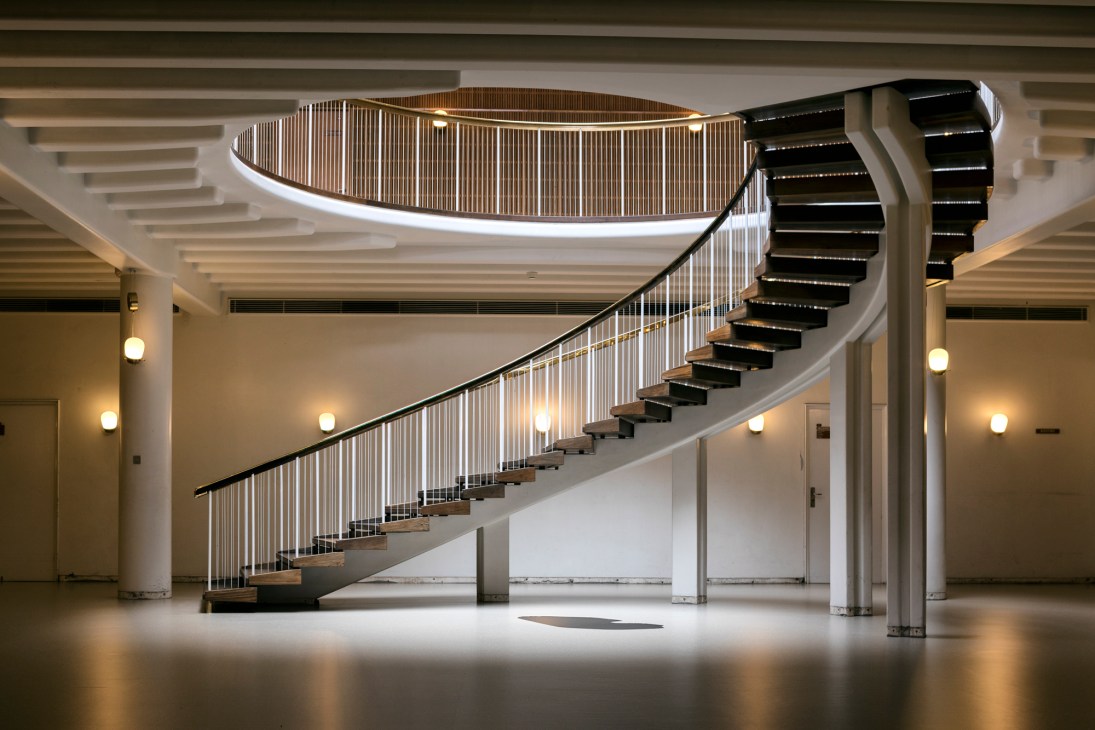 A modernist spiral staircase with metal railings and wooden steps curves upward in a bright atrium with recessed lighting.