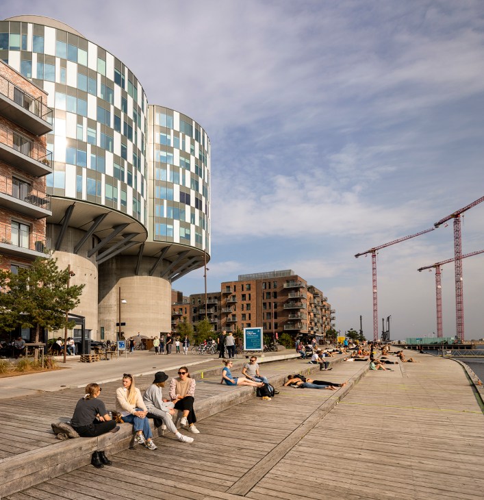 Modern waterfront development with contemporary residential buildings, wooden decking, and people relaxing outdoors