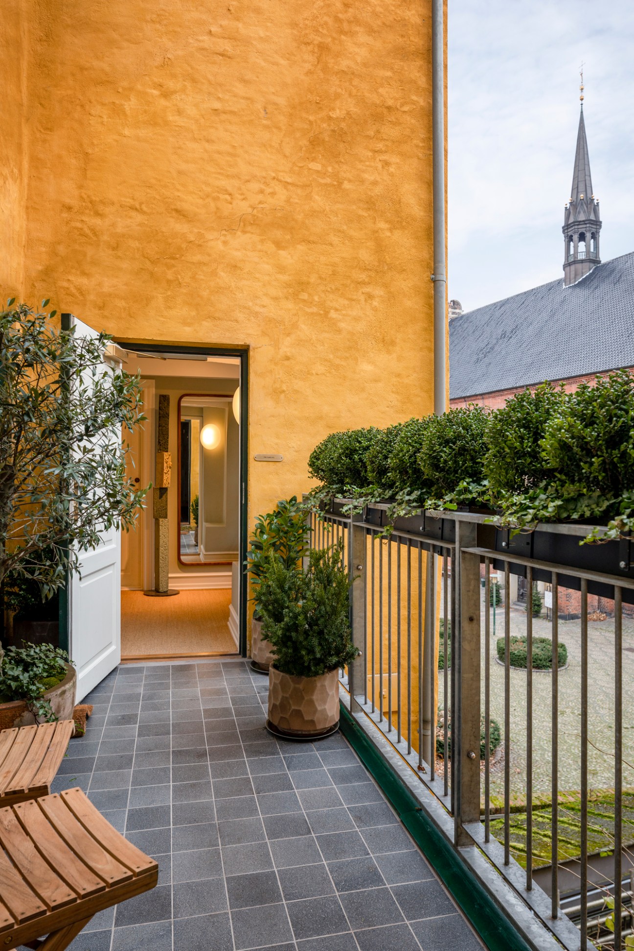 Private terrace entrance to The Darling hotel with yellow ochre facade, potted plants, and Copenhagen church spire in background.