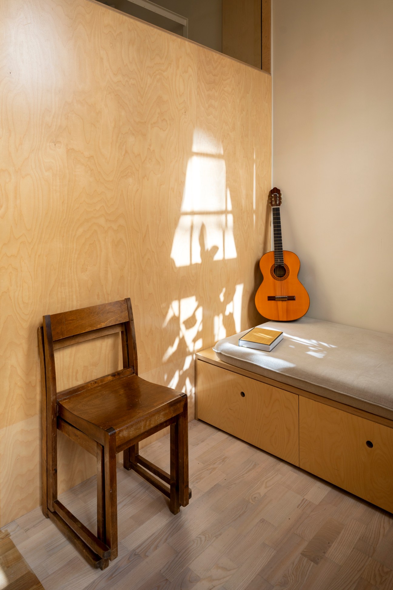 Minimalist interior with light wood walls, acoustic guitar, wooden chair, books, and window casting geometric shadows.