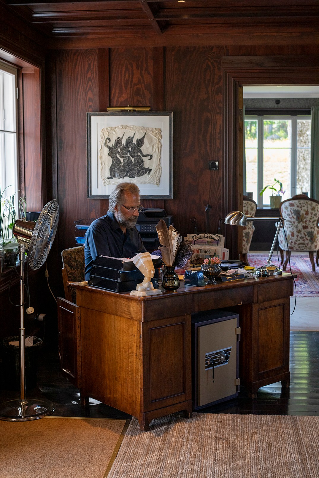 Man with beard and glasses reading at wooden desk in wood-paneled study with quill pen and vintage decor
