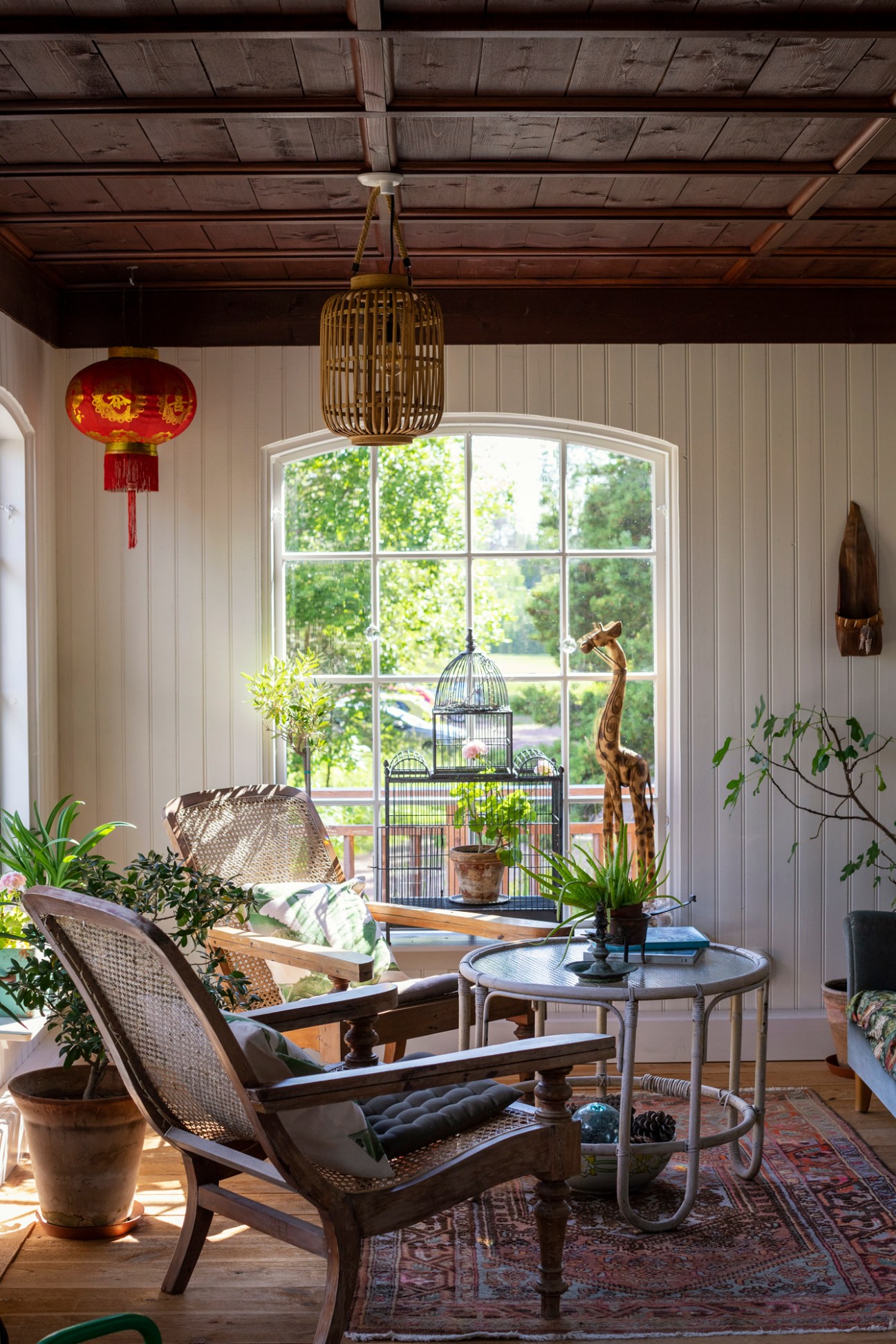 Sunlit interior room with wicker chairs, wooden ceiling beams, large arched window, birdcage and plants