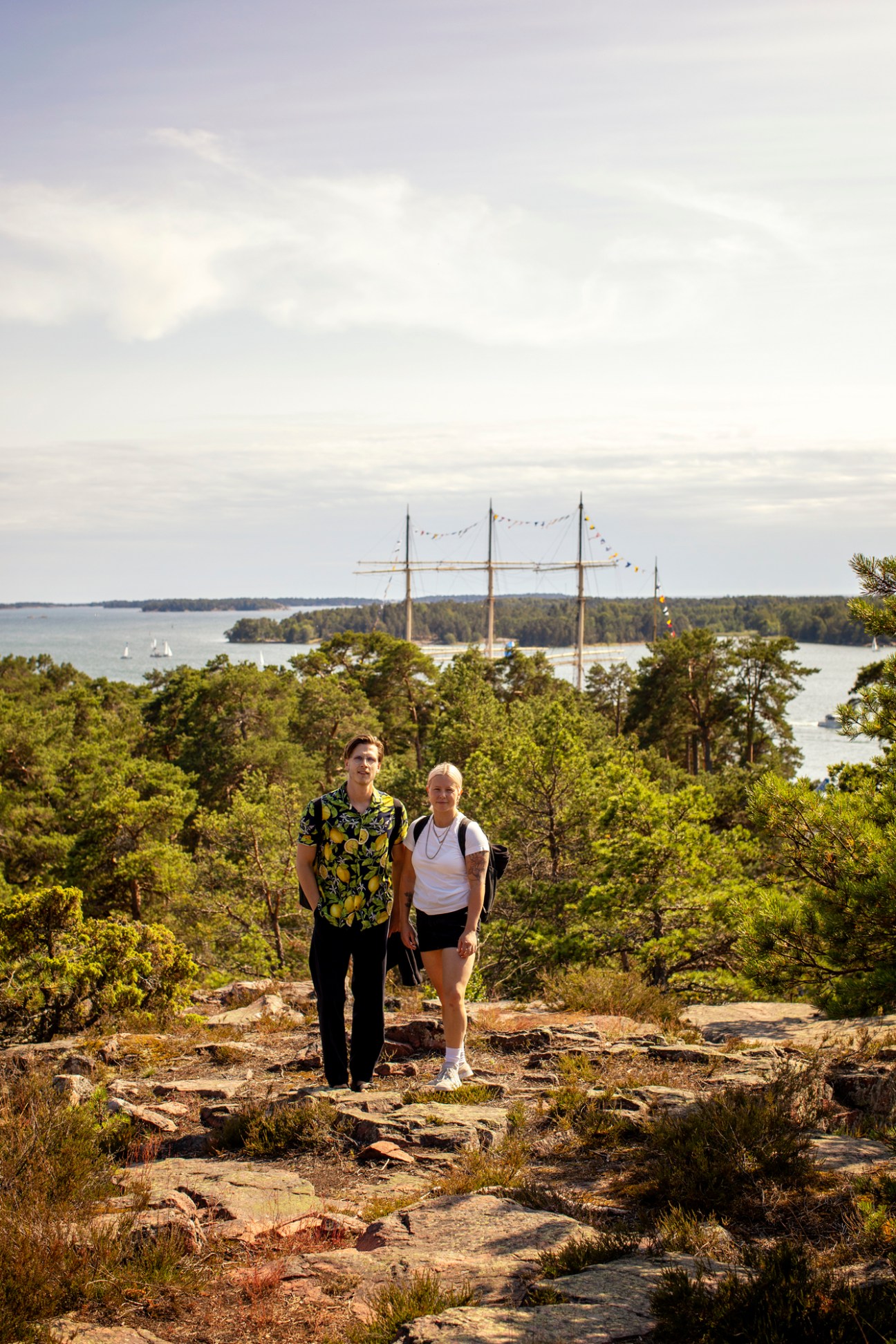 Two people standing on rocky coastal terrain overlooking water with tall-masted sailing ship in background