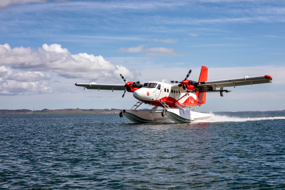 Red and white Twin Otter seaplane landing on water with coastal landscape in background.