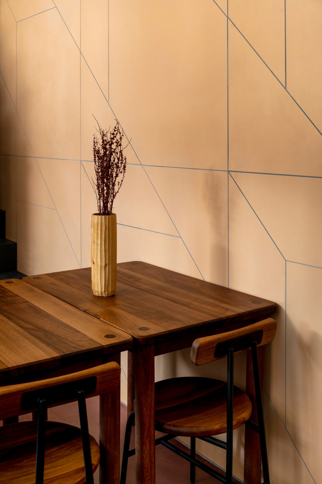 Restaurant interior with wooden high table, stools, and dried plant arrangement against geometric wall pattern