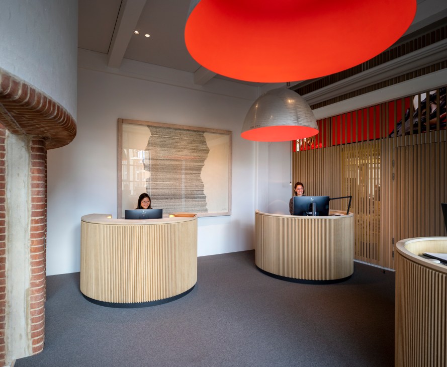 Modern reception area with two circular wooden desks and staff, red overhead lighting, and natural materials in Lindberg's Aarhus headquarters.