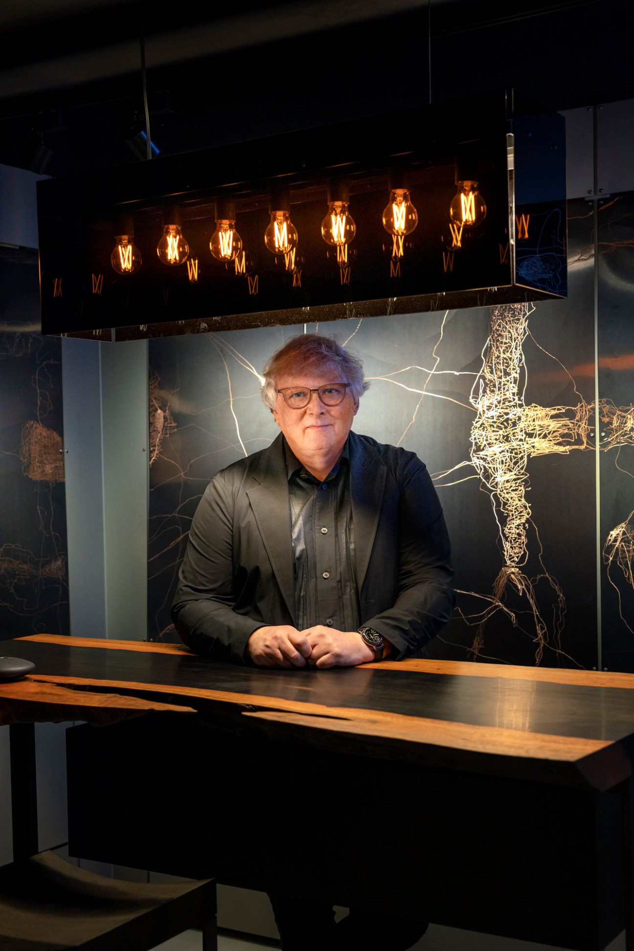 Henrik Lindberg, CEO of Lindberg eyewear, stands at a desk beneath illuminated lightbulbs and abstract wire art installations.