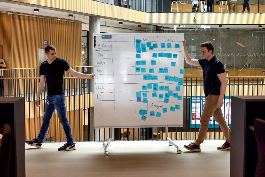 Two men move a mobile whiteboard covered in blue sticky notes through a modern office space with wooden features