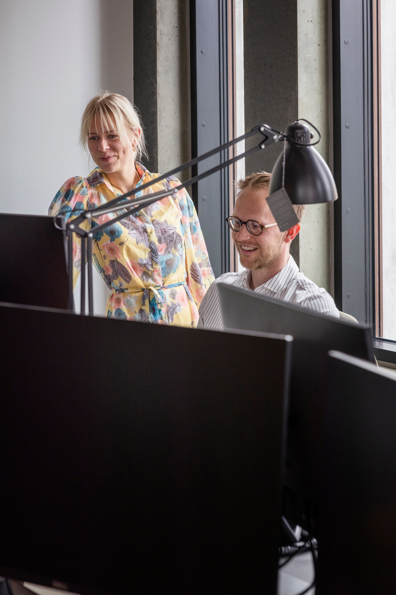 Two journalists collaborating at desks in open newsroom with woman standing and man sitting at computer