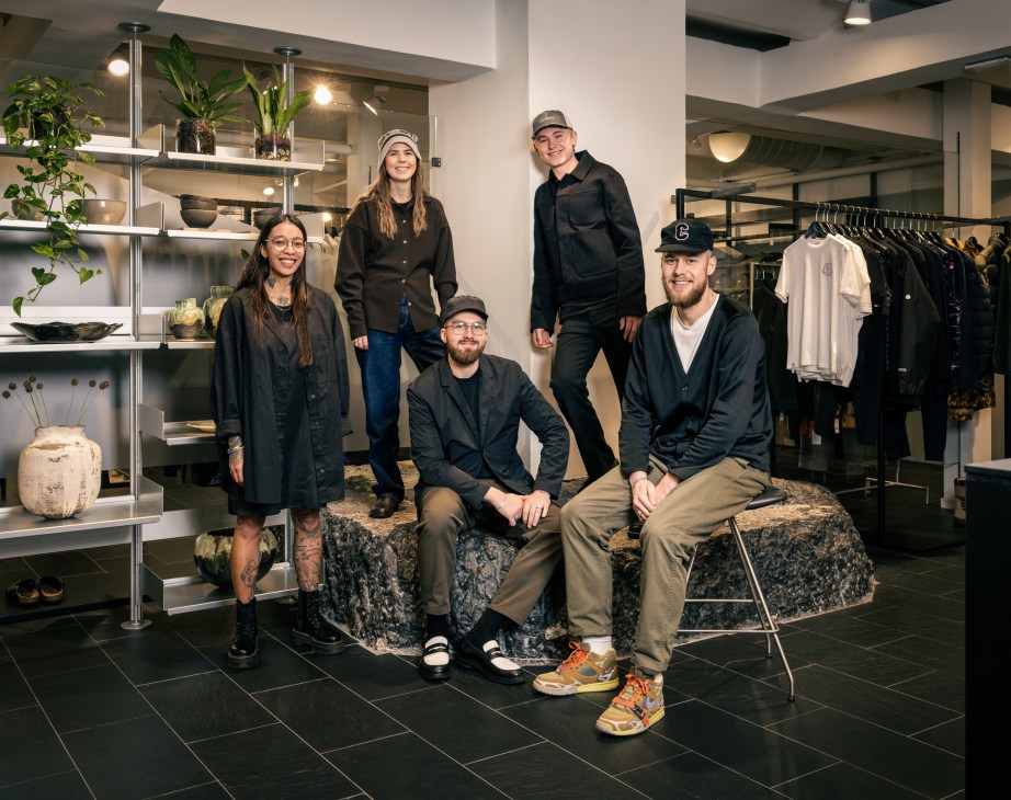 Five team members posing together in a modern menswear retail store interior with black and white clothing displays, shelving with plants and decorative items, and clothing racks visible in the background.