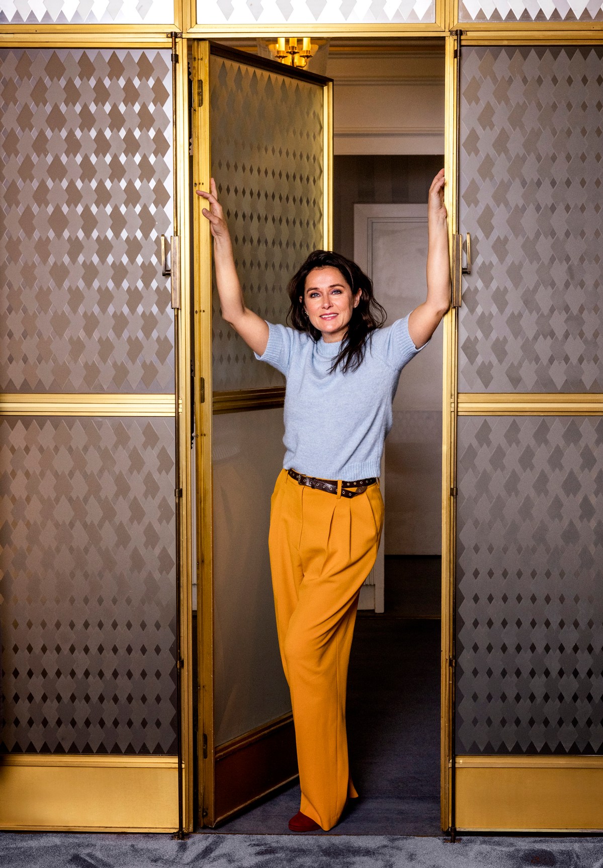 Sidse Babett Knudsen standing in doorway of ornate gold-framed glass doors with geometric patterned walls