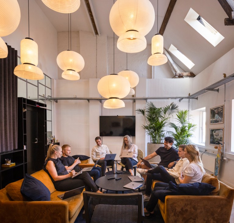 Podimo founders and team members sit in a modern office lounge with camel-brown sofas and hanging pendant lights.