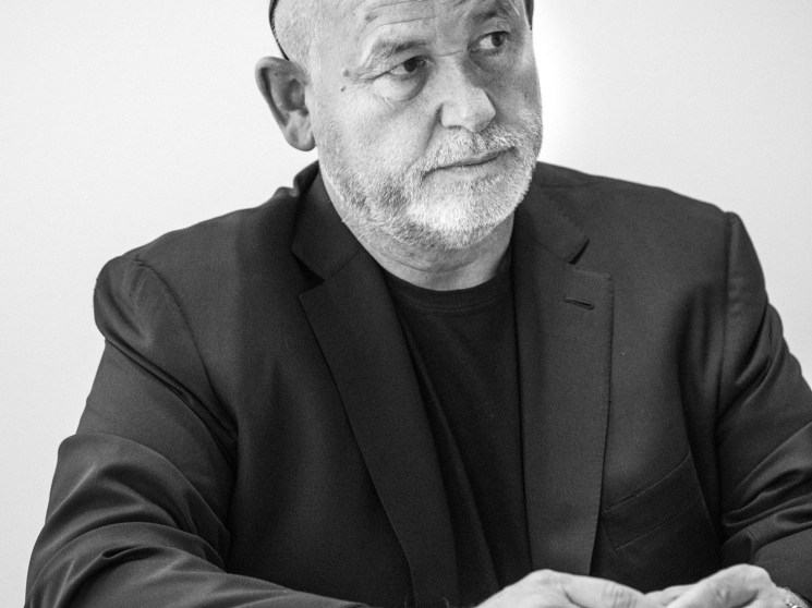 Black and white portrait of an older man in a blazer with glasses perched on his head, seated at a table in thought.