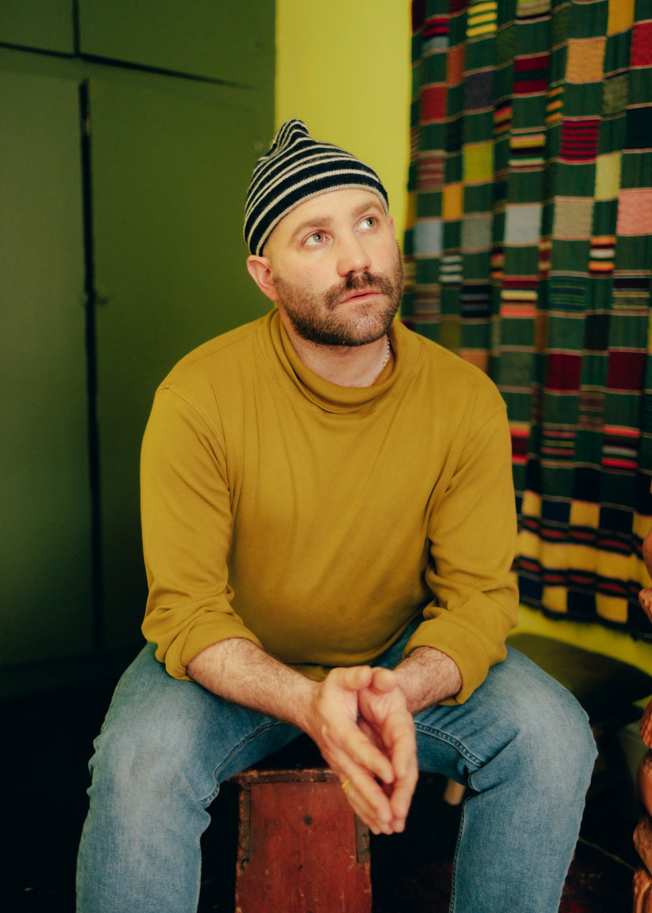 Man in yellow sweater and striped beanie sitting against green wall with patterned textile backdrop.