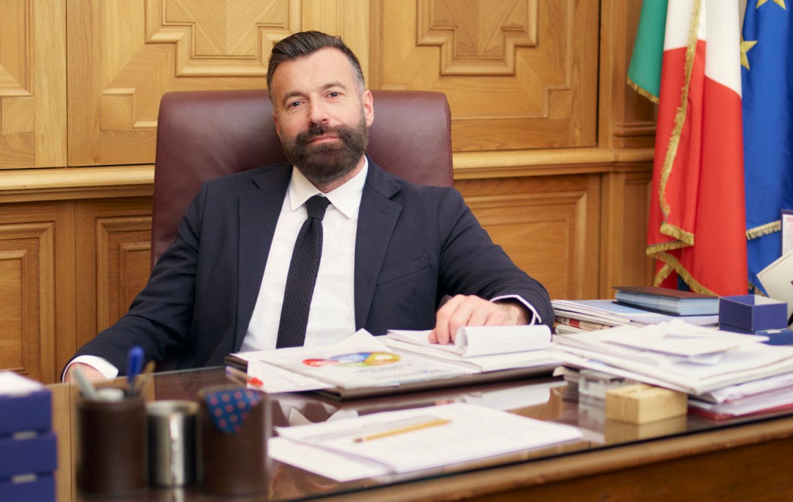 Alessandro Zan sitting at his desk in an ornate office with wood paneling and flags