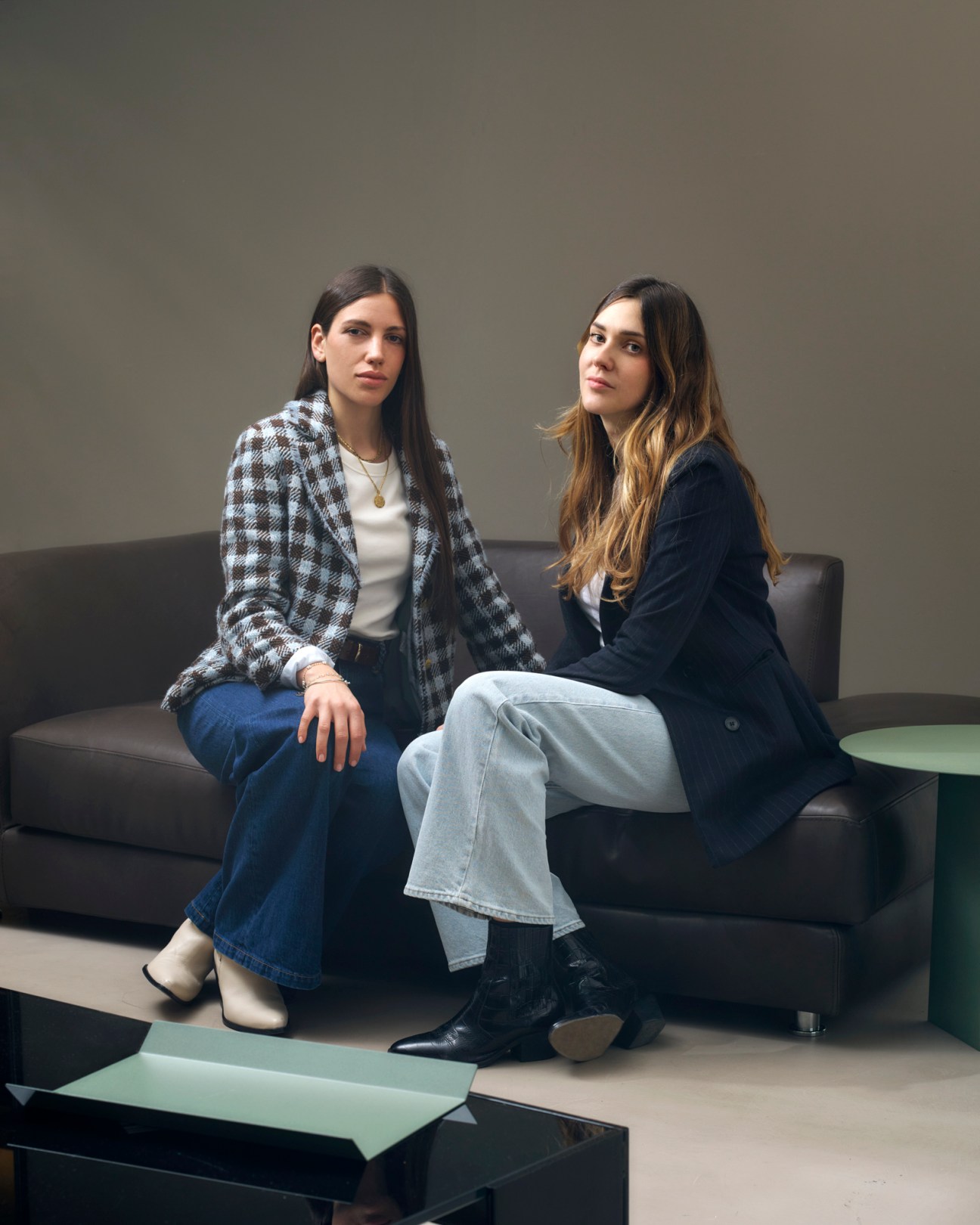 Two women sitting on dark sofa, one wearing checkered blazer and jeans, other in navy blazer and light jeans