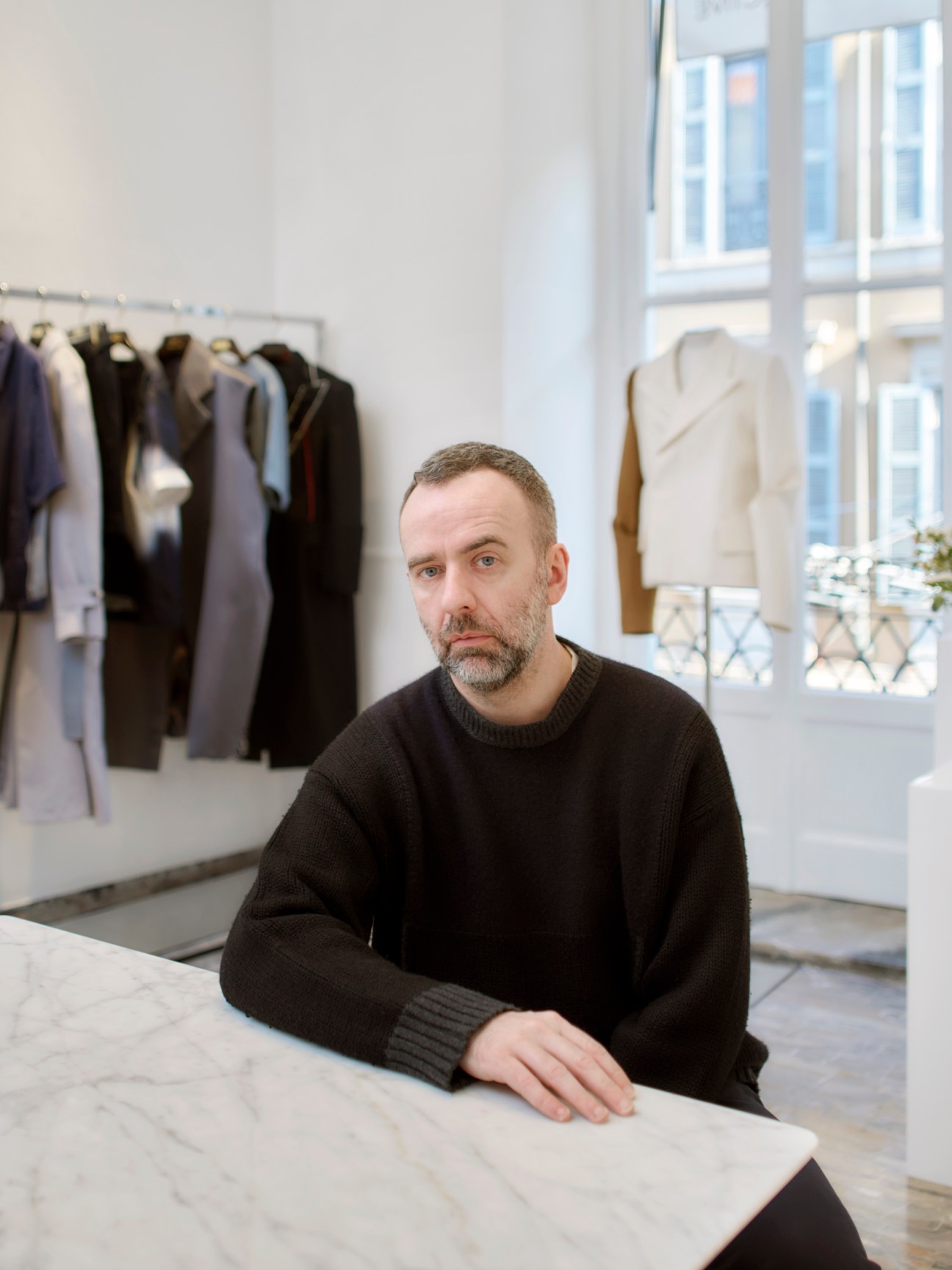 Man in black sweater standing at counter in modern showroom with clothing displayed on wall behind him
