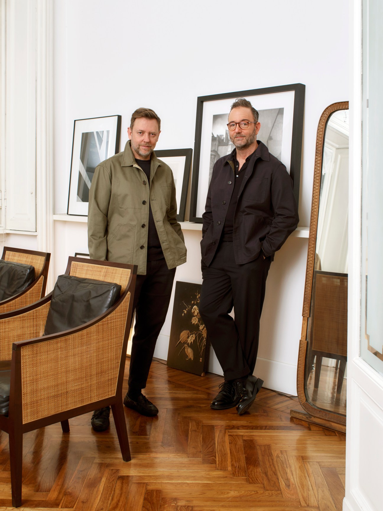 Two men standing in a minimalist interior space with framed artwork and rattan chairs