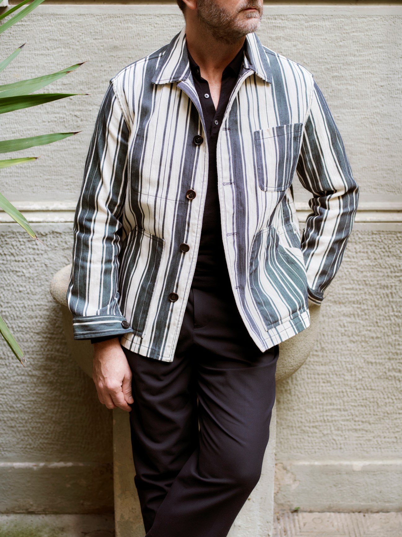 Man wearing a striped cotton twill jacket over black trousers against a concrete wall with green foliage.
