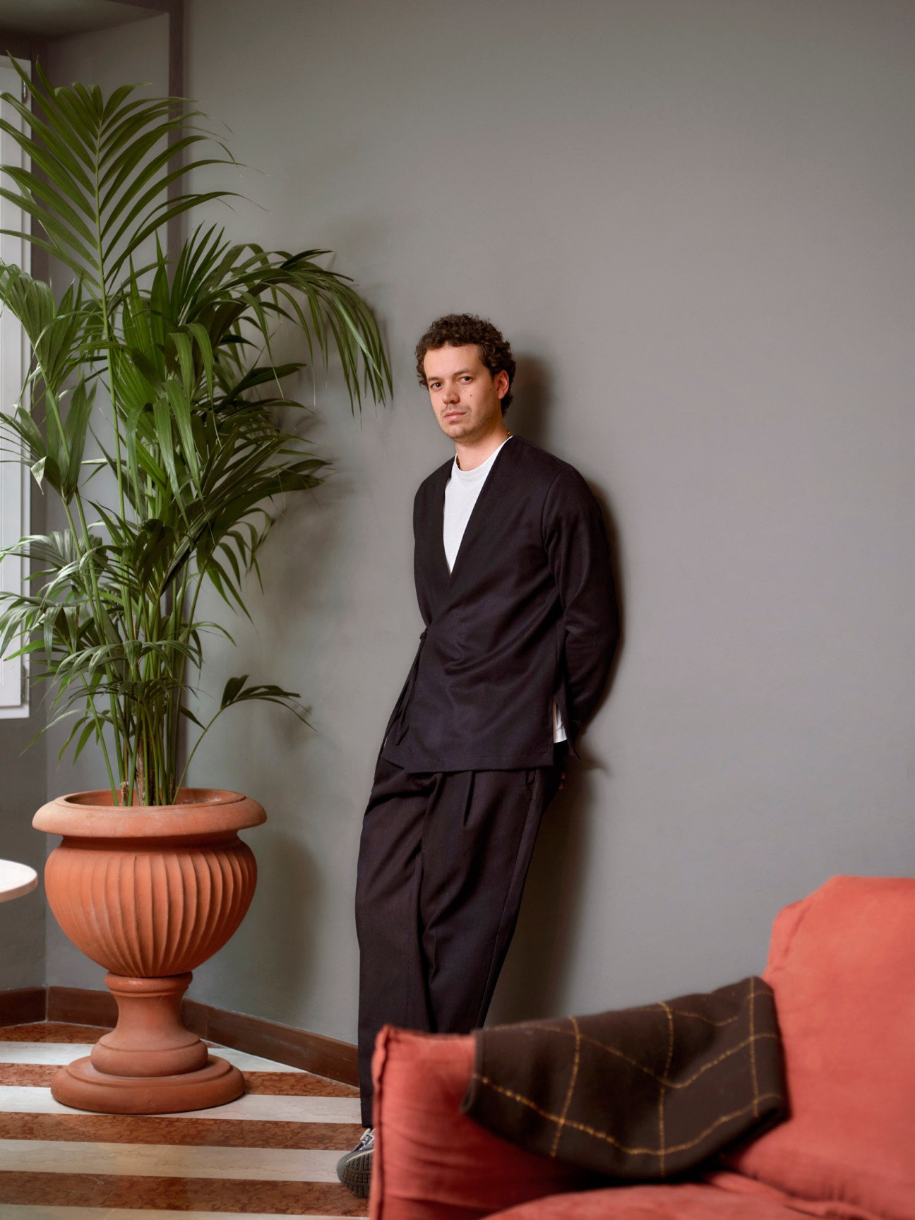 Man wearing black blazer and trousers standing in minimalist interior with potted plant