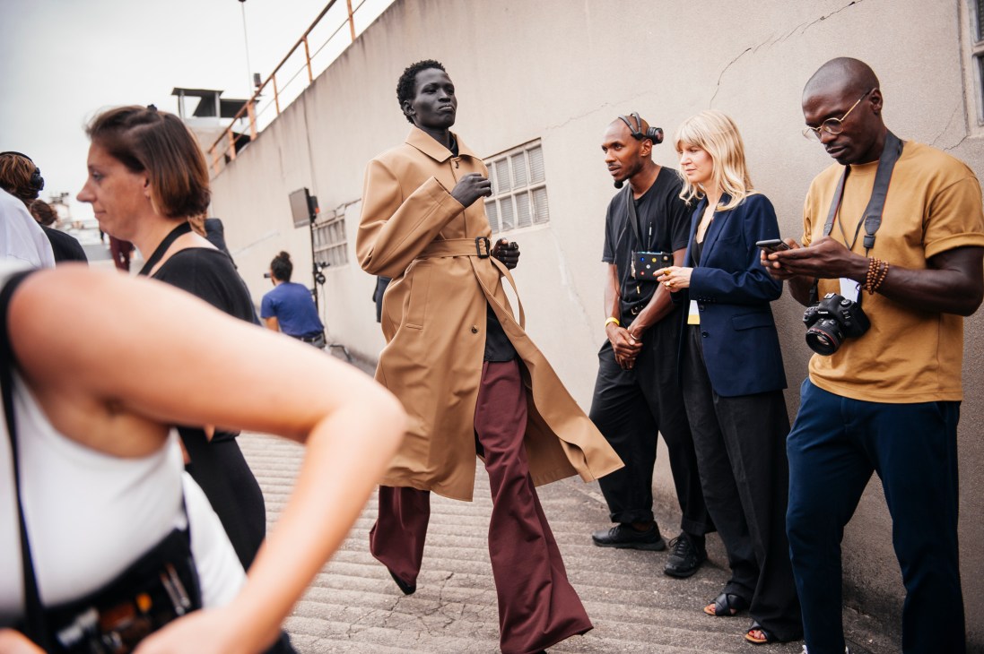 Model in tan trench coat and burgundy trousers walking past photographers and crew backstage at fashion show