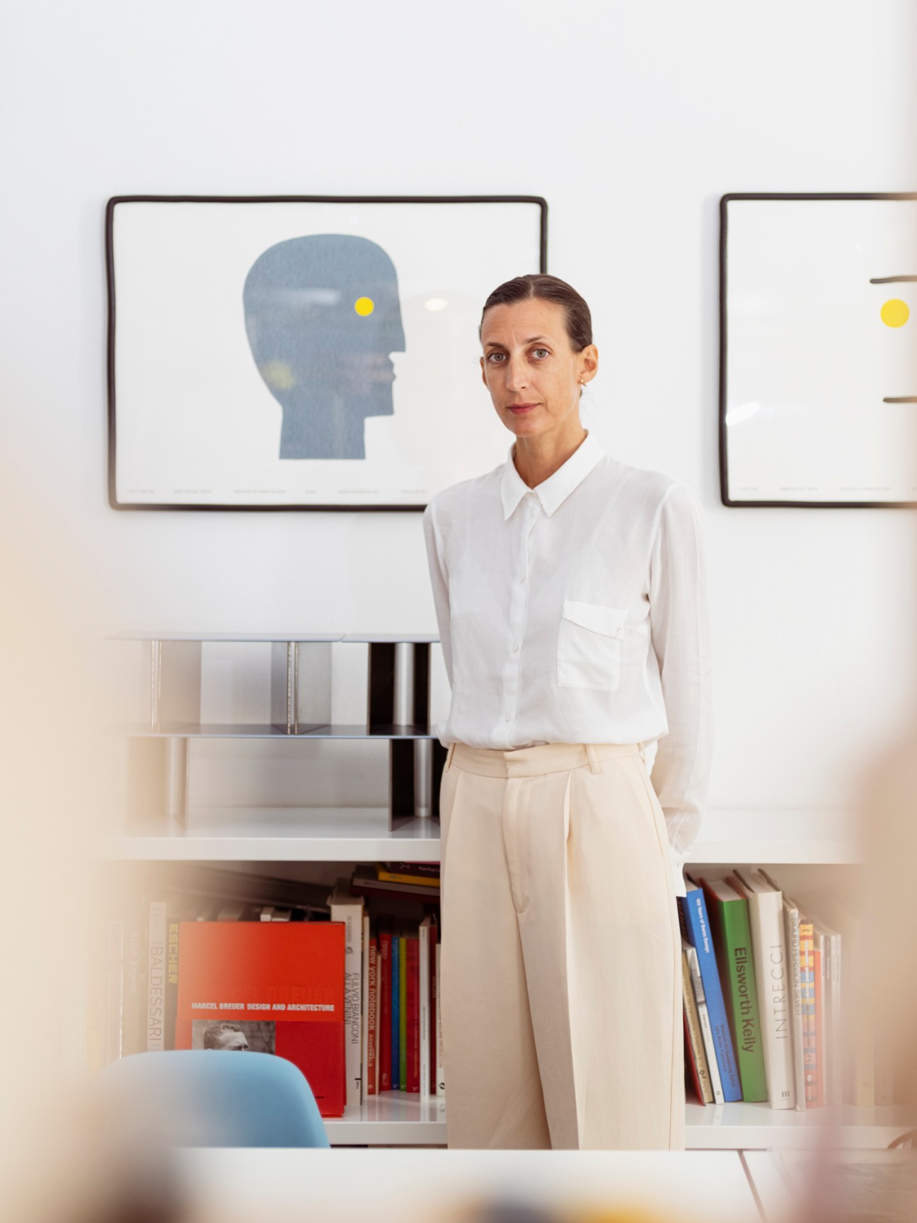 Woman in white shirt and cream pants standing in modern interior with framed artwork and built-in shelving