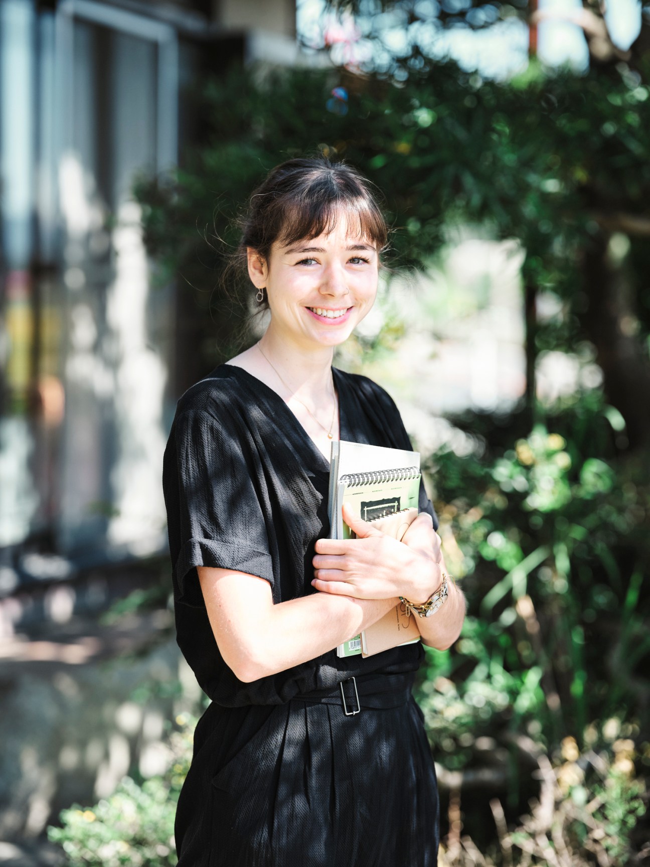 Young woman in black dress holding notebook, smiling outdoors in garden setting.