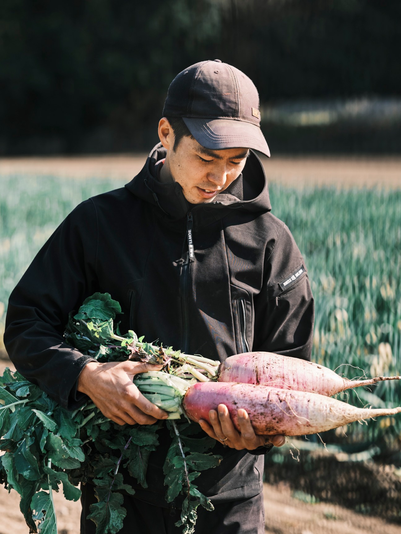 Young farmer Ryohei Tanaka holding fresh root vegetables in a field wearing dark clothing and cap