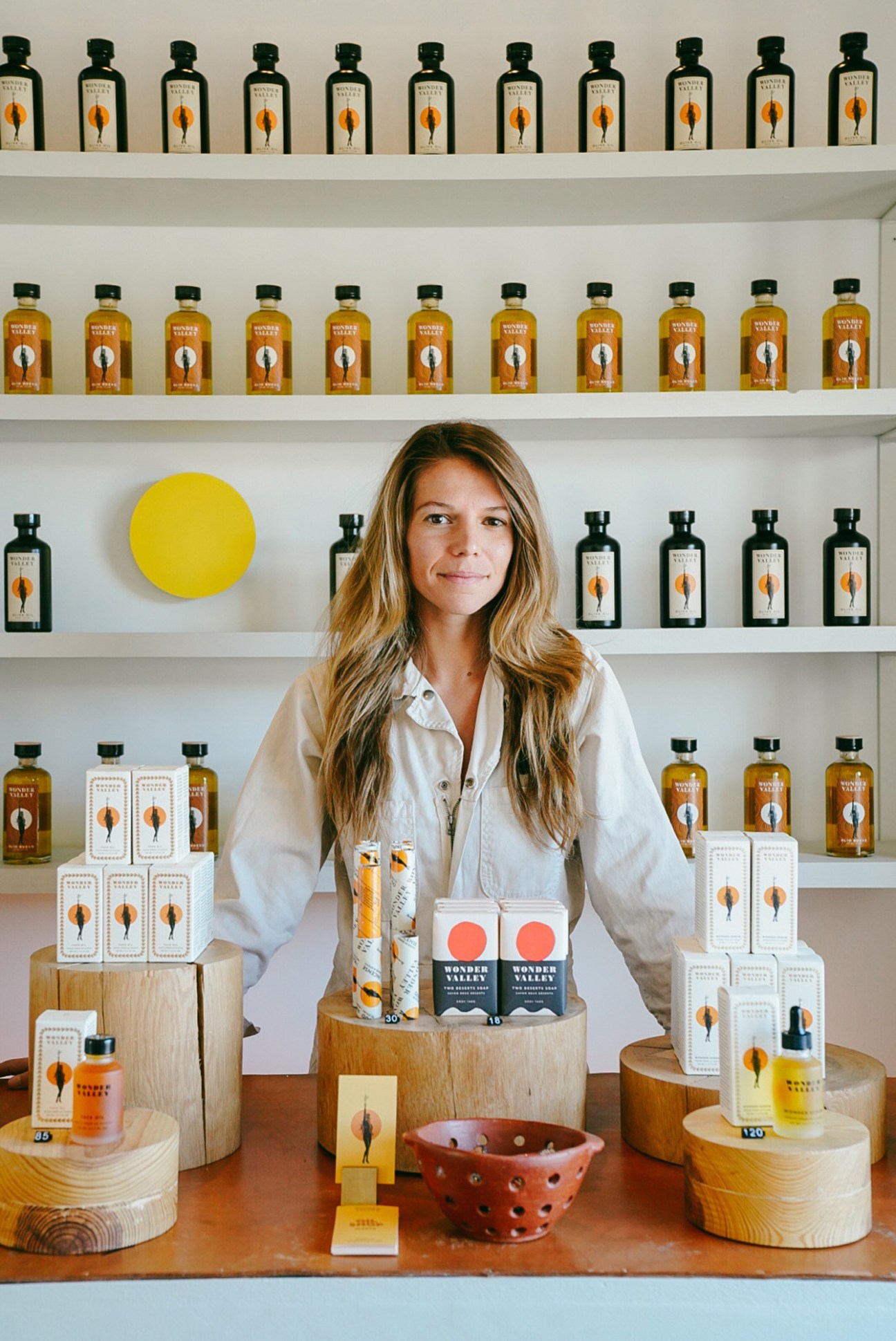 Woman with long blonde hair in white shirt standing behind display of Wonder Valley olive oil products on wooden stands