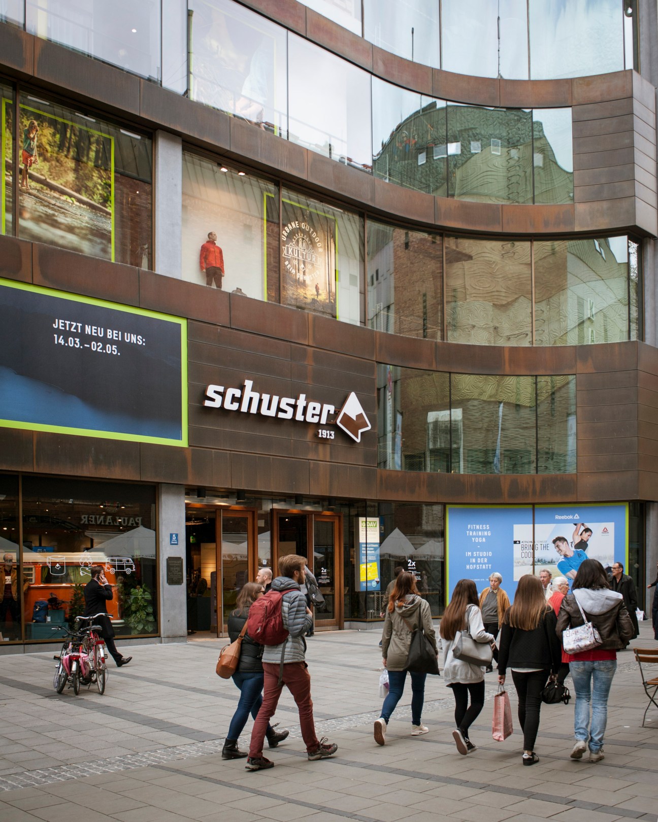 Sporthaus Schuster storefront in Munich with curved glass facade, brown metal cladding, and pedestrians walking past.