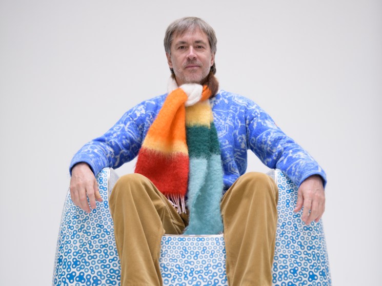 Man with beard sitting on blue patterned bean bag chair wearing colorful striped scarf and blue sweater